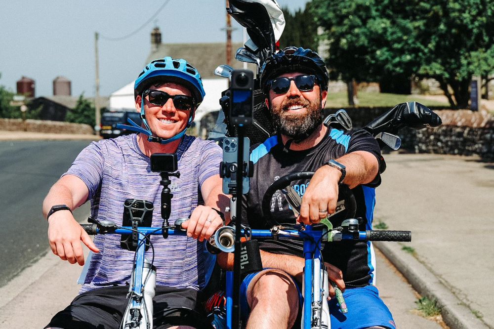 Two men sitting side by side on bikes wearing Prostate Cancer UK cycling gear and sunglasses, smiling