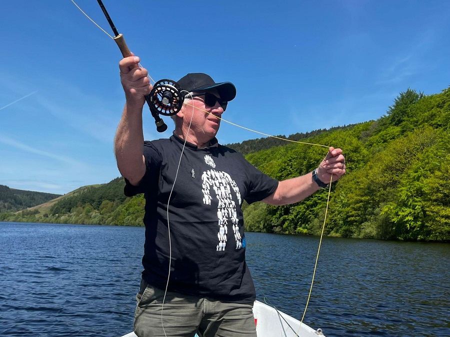 A man wearing a Prostate Cancer UK tee-shirt standing on a boat on water, holding a fishing line
