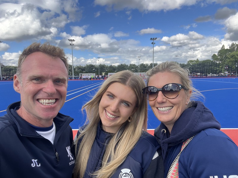 A man in a blue sports jacket taking a selfie with his wife and teenage daughter by the side of a hockey pitch.