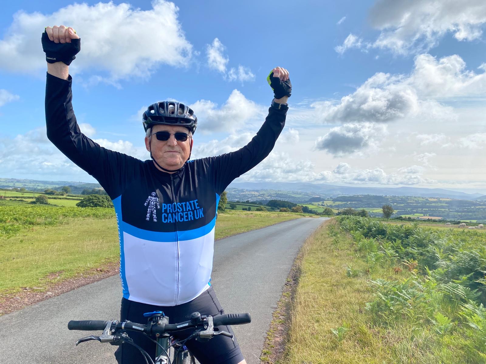 A man wearing Prostate Cancer UK branded cycling uniform stands astride a bicycle with his hands raised in celebration.