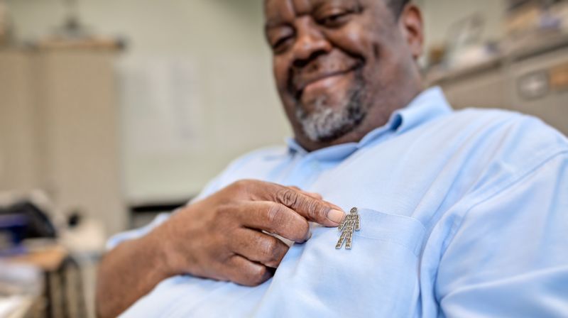 A man wearing a blue shirt pointing to a Man of Men logo badge on his chest. 