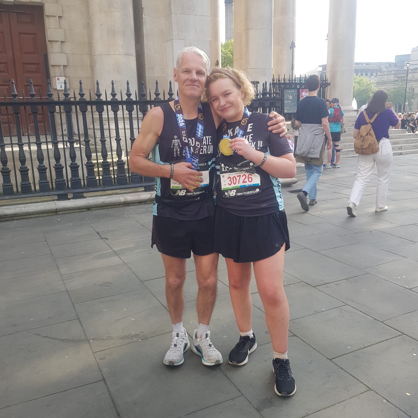 A man and a young woman wearing black Prostate Cancer UK running kit and holding their Marathon medals.