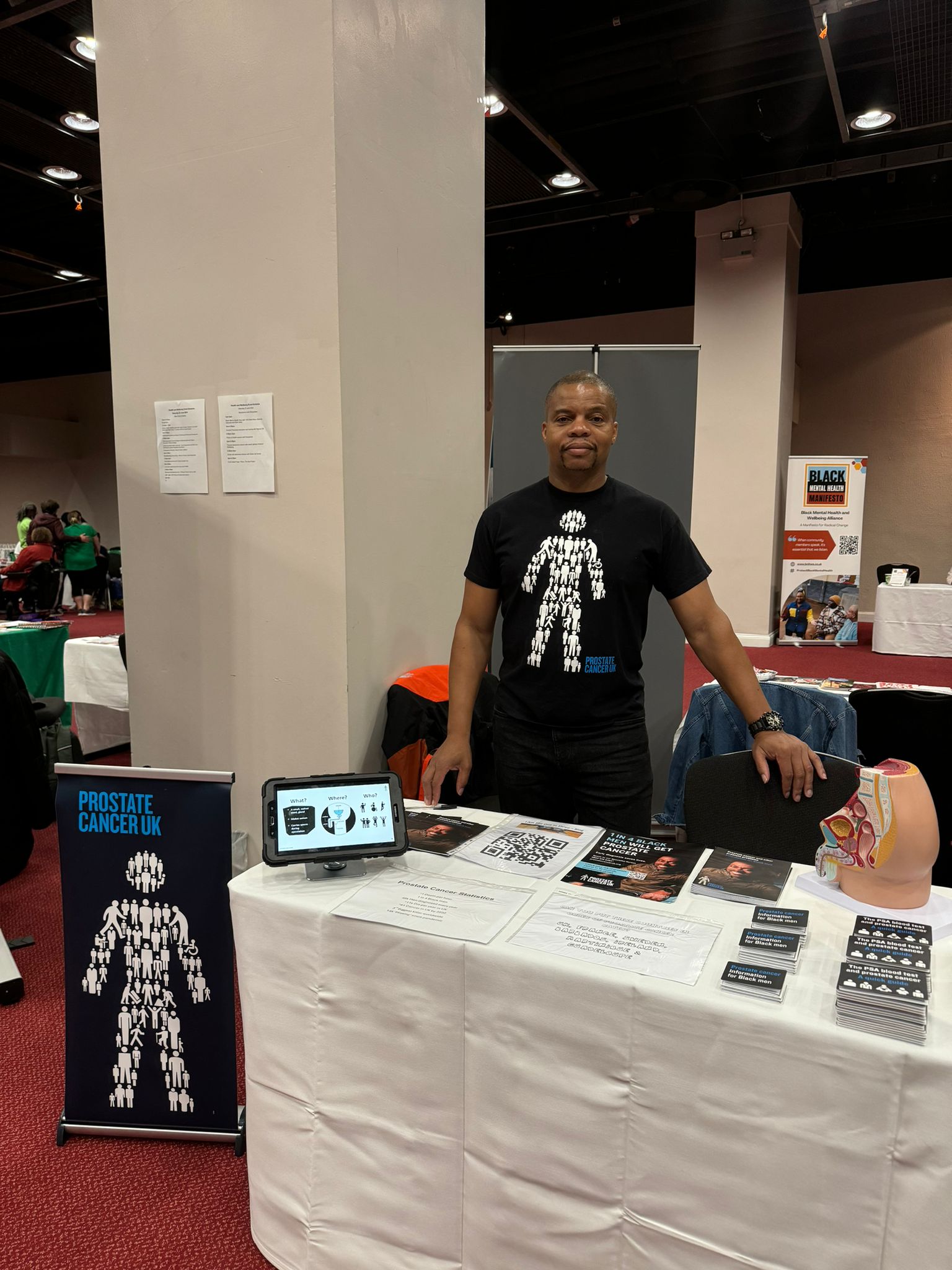 A man wearing a black Prostate Cancer UK t-shirt hosting an awareness stand and smiling at the camera.