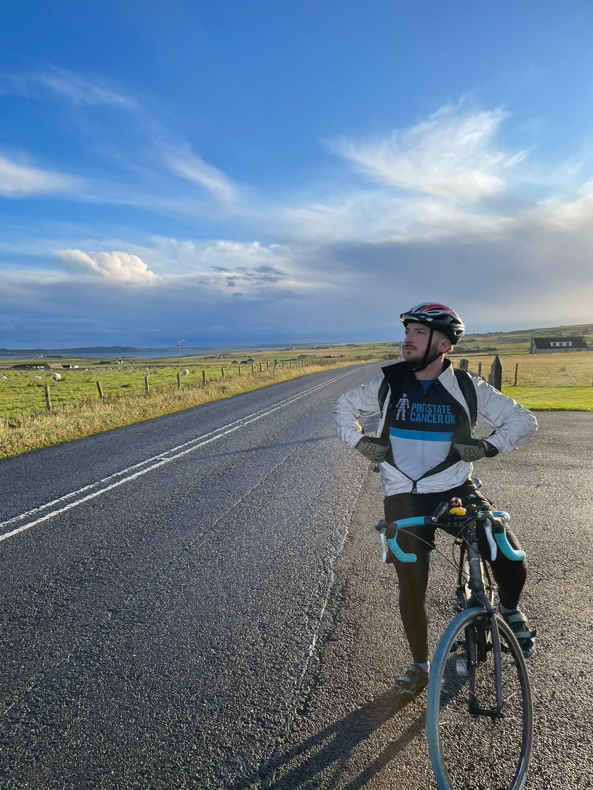 A man on a bike wearing Prostate Cancer UK cycling gear, on a road surrounded by fields