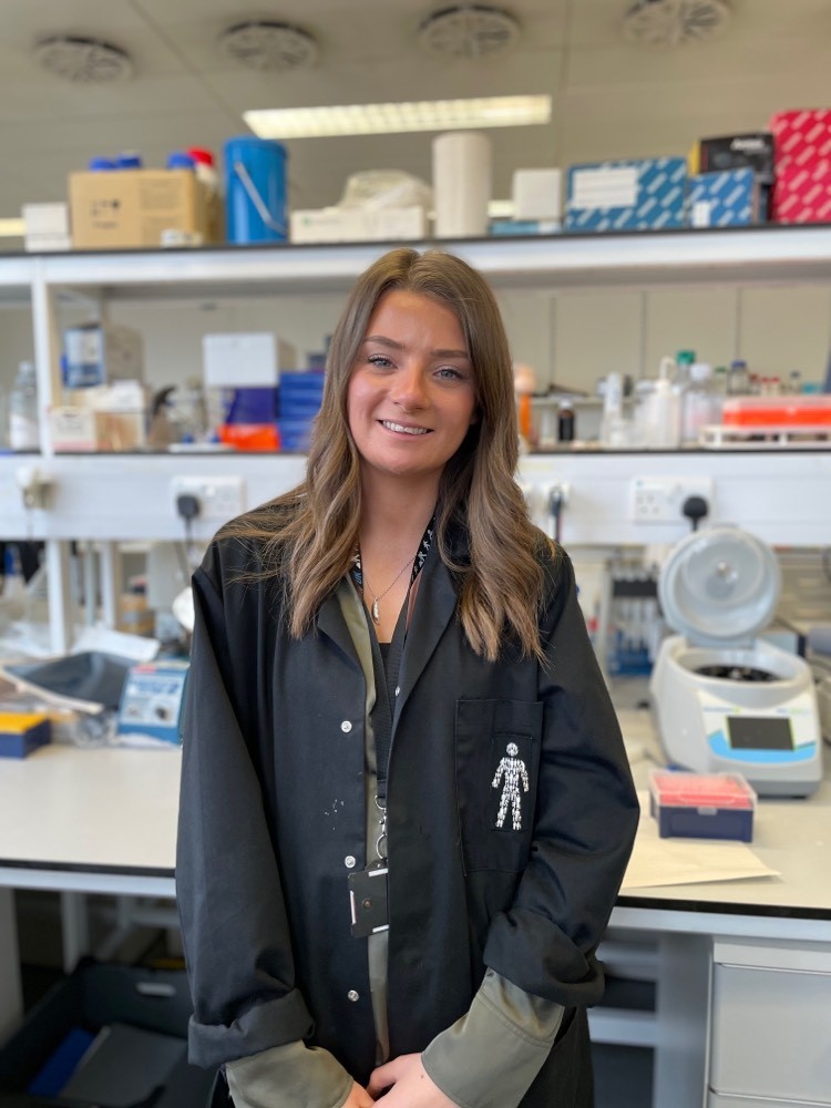 A woman with mid-length hair wearing a dark blue jacket with a Prostate Cancer UK badge pinned on, standing in a lab and smiling