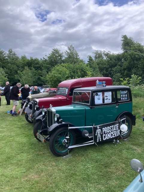 A line of classic cars parked on some grass, with a Prostate Cancer UK banner next to them
