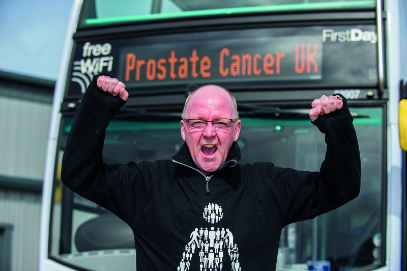 A man wearing a Prostate Cancer UK sweater punches the air in celebration while standing in front of a FirstGroup bus.