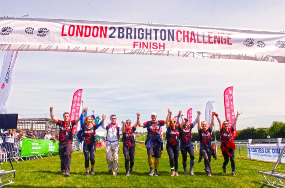 A group of people at the London2Brighton Challenge finish line, standing in a line with raised hands