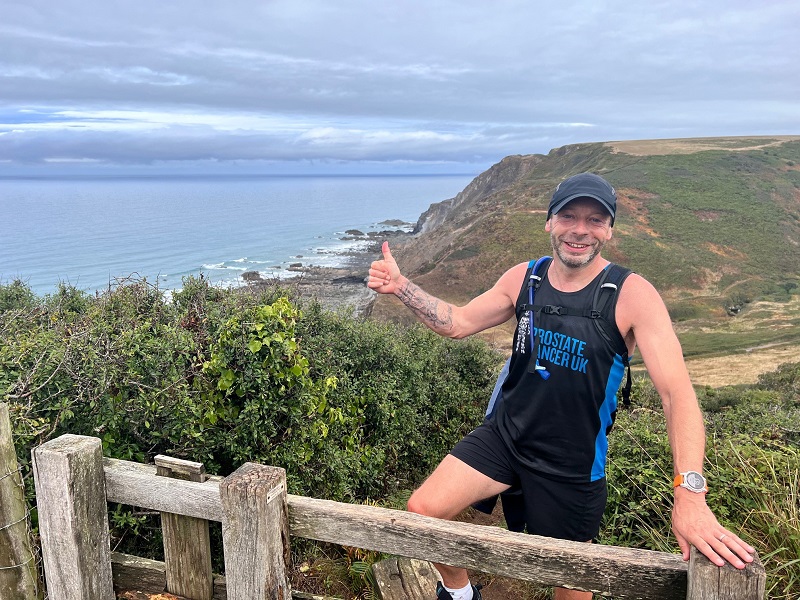 A man wearing a Prostate Cancer UK vest standing at a stile on a coastal path, giving a thumbs up
