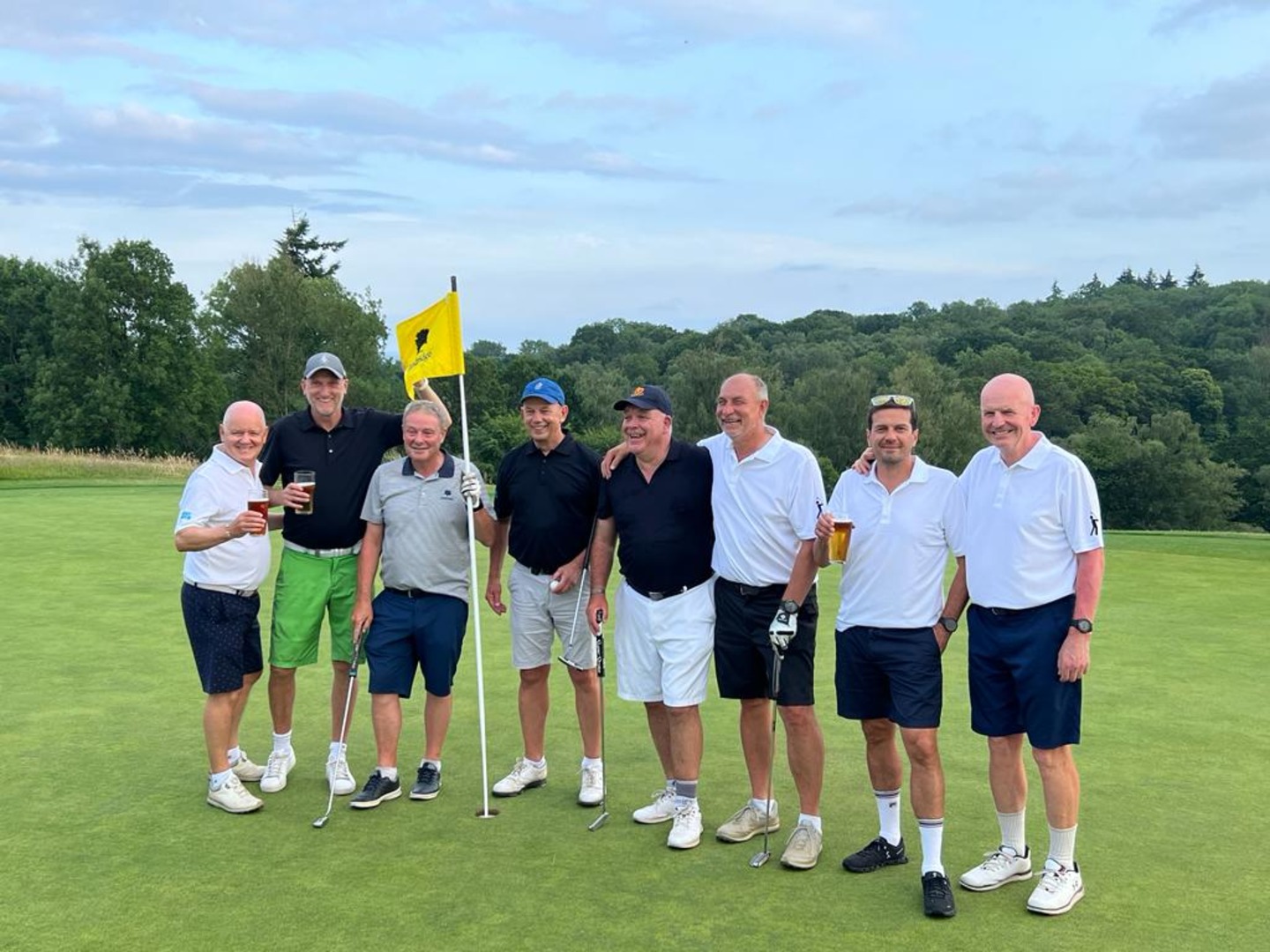 A group of eight men wearing golf uniform pose for a photo while standing next to a flag on a golf course green