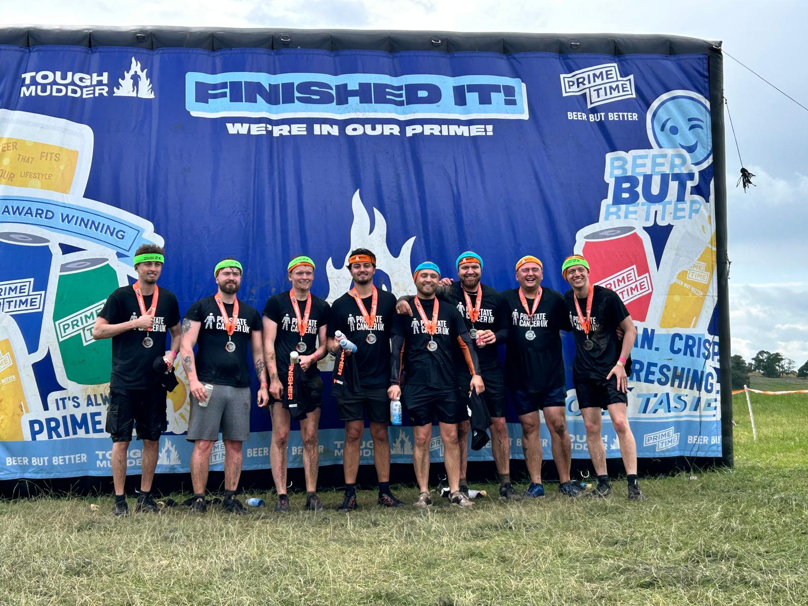 A group of men wearing shorts and t-shirts, standing by a Tough Mudder finish banner with their medals