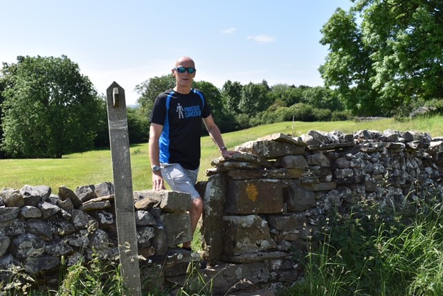 A man wearing a Prostate Cancer UK tee-shirt walking through a gap in a stone wall by a field on a sunny day