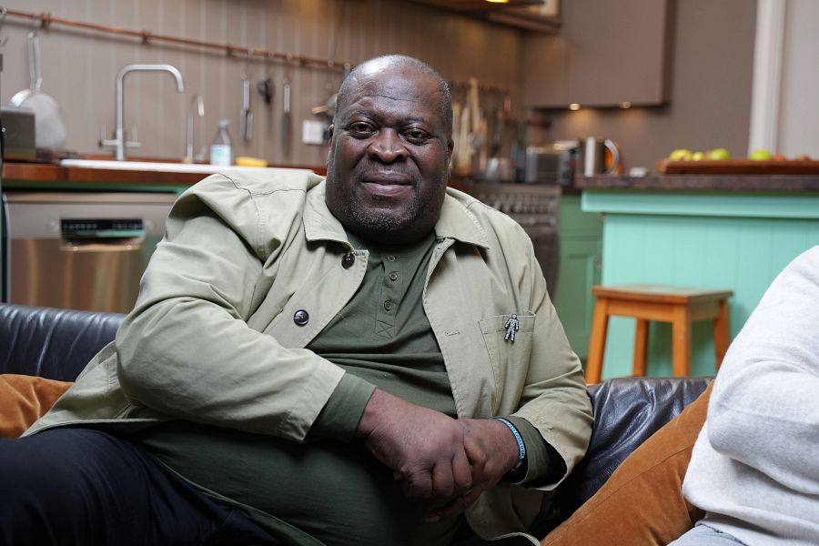 A Black man wearing a beige jacket and green t-shirt, smiling while sitting on a sofa.
