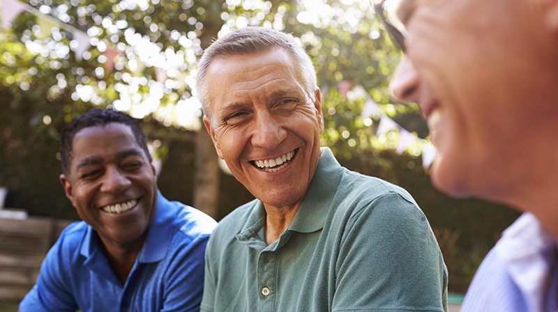 Three men sat in a garden smiling and talking with each other.