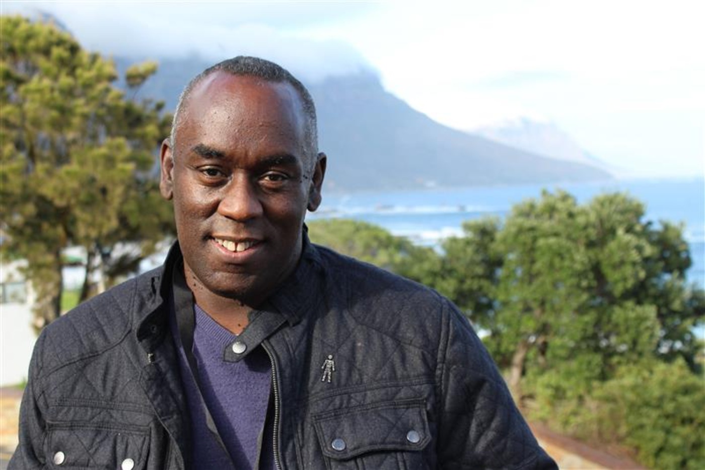 A man wearing a black jacket and a Prostate Cancer UK 'Man of Men' badge stood in front of mountaintop scene