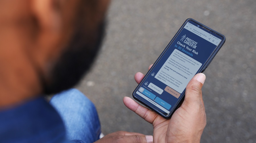 A man holding a mobile phone showing the Prostate Cancer UK risk checker.