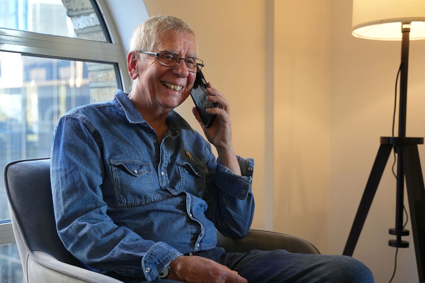 A man wearing a blue denim shirt with a Prostate Cancer UK Man of Men badge smiles while talking on the phone in a living room