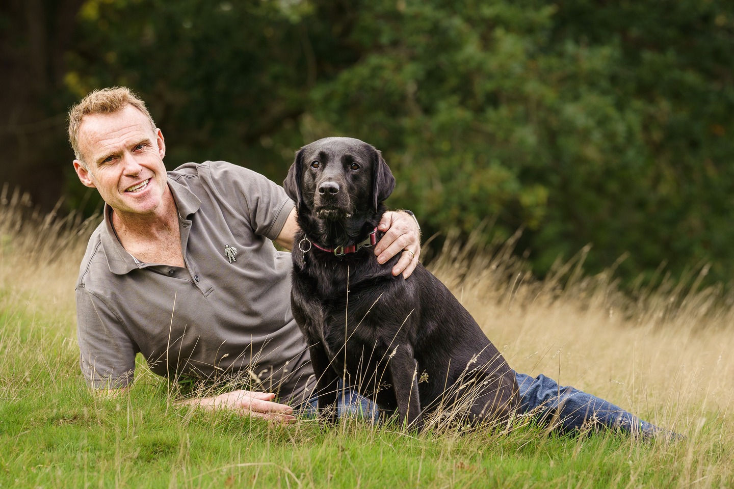 A man wearing a grey shirt lying in a grassy field with his arm around a Black Labrador dog