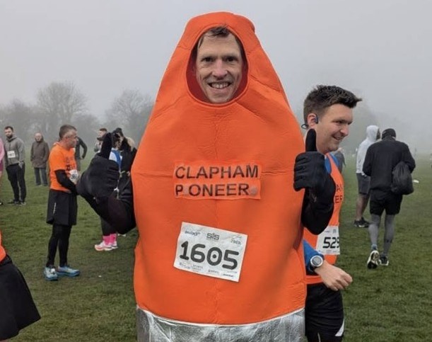 A man smiling and putting his thumbs up while wearing an orange traffic cone costume at the start of a running event.
