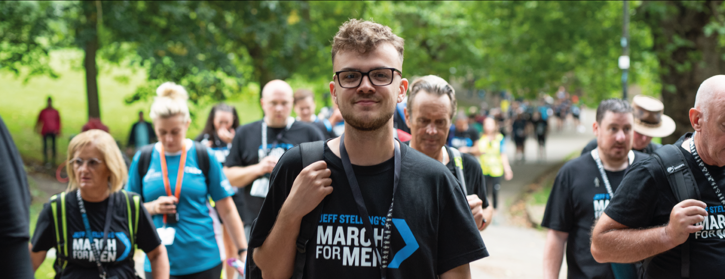 A man wearing a charity smiles while taking part in a sponsored walk event in a park.