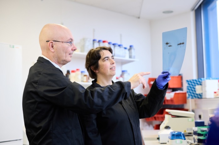A man wearing as suit standing with a woman wearing a dark coat in a lab, looking at a graph on paper