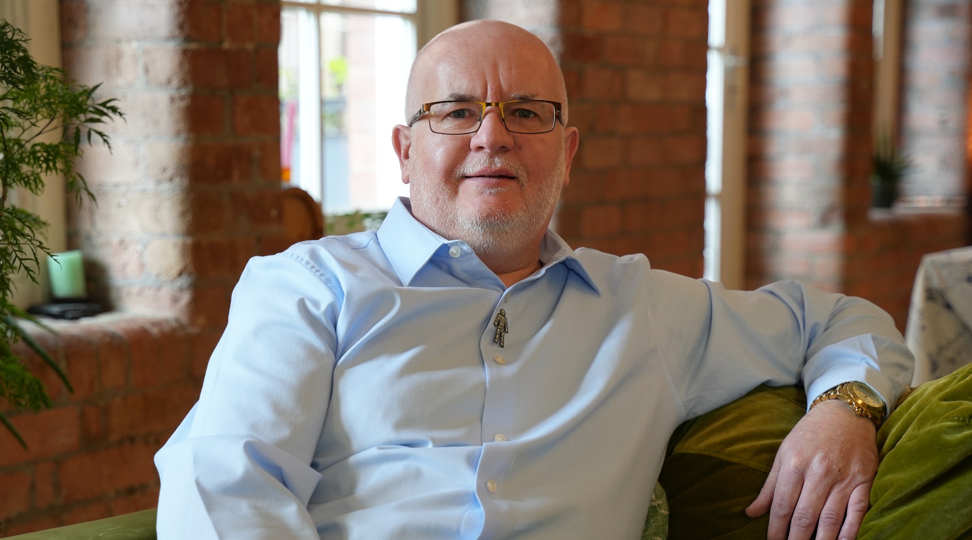 A man wearing a blue shirt leaning back in an arm chair with his arm rested to the side.