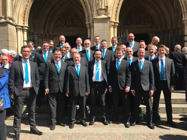 A male choir wearing suits and blue ties standing in a group outside a church