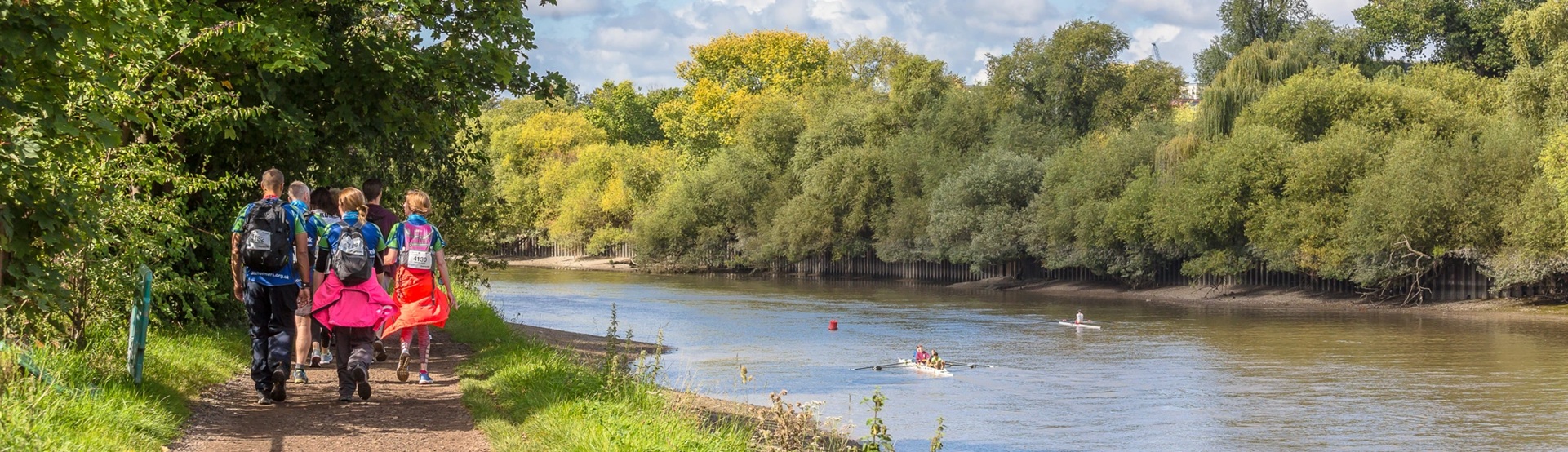 A group of adults walking along a tree-lined pat by a river, where people are rowing