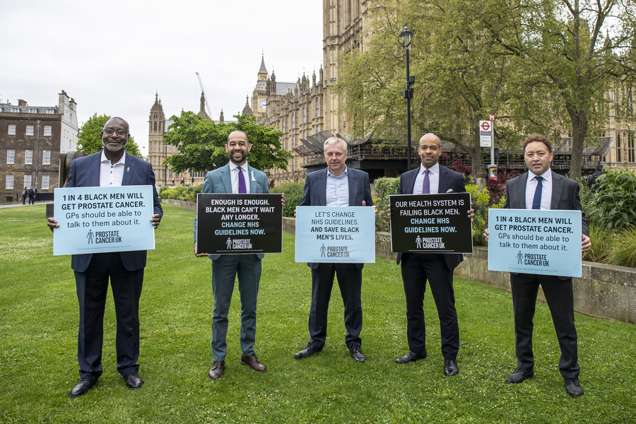 Five men wearing suits stand shoulder to shoulder holding up large placards displaying information about prostate cancer.