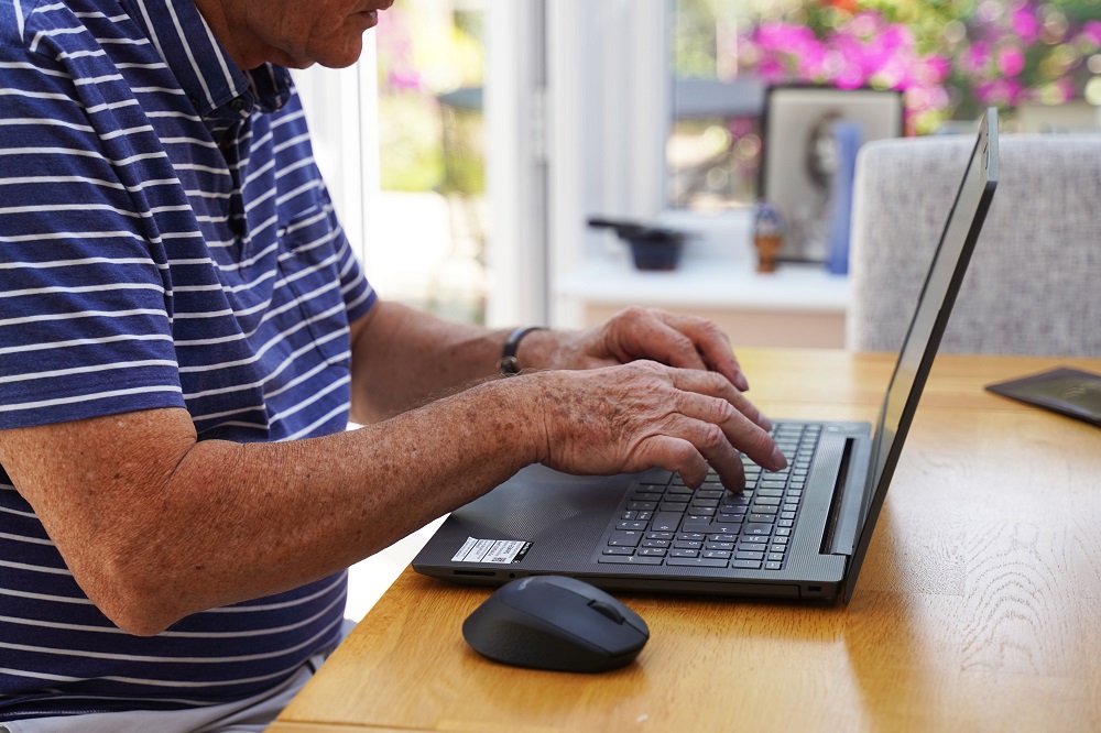 A man wearing a blue shirt sitting at a table and typing on a computer. 
