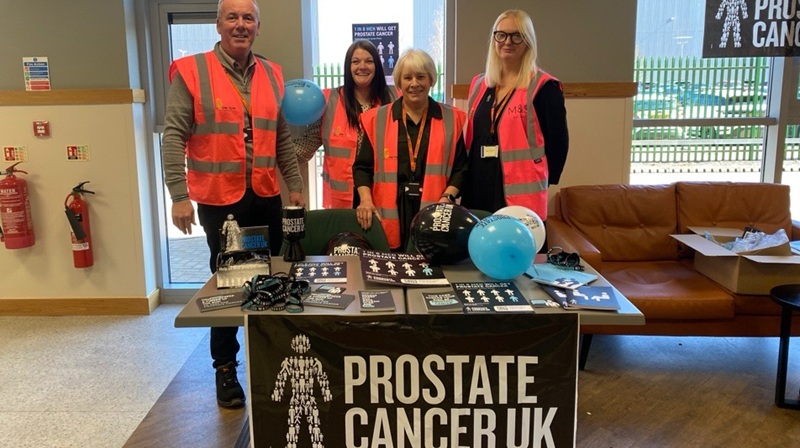 Four people wearing high vis jackets at a Prostate Cancer UK fundraising stand with leaflets and balloons