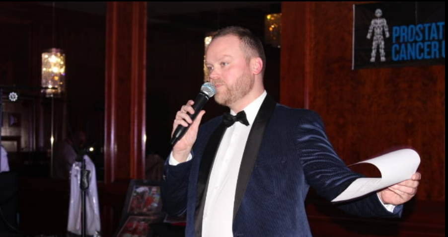 A man wearing a suit and bow tie talking into a microphone and holding a piece of paper