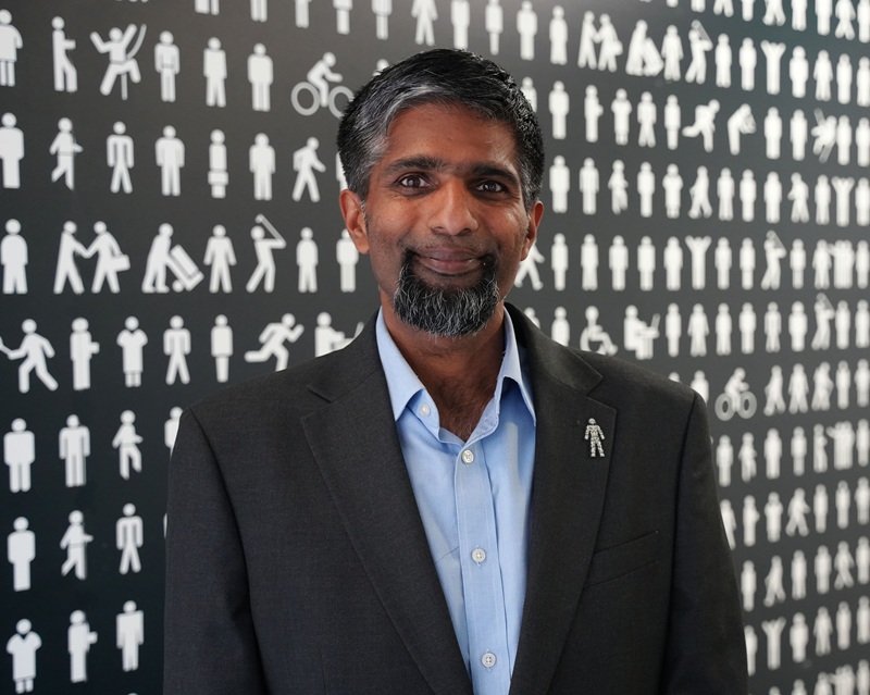 Rajit Gholap wearing a black jacket and blue shirt standing in front of the Prostate Cancer UK backdrop