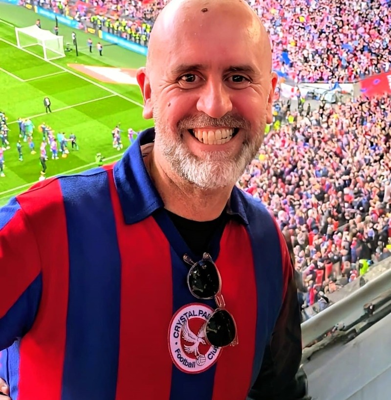 A man wearing a red and blue striped replica football shirt smiles while standing amongst a huge crowd of fans at Wembley Stadium.