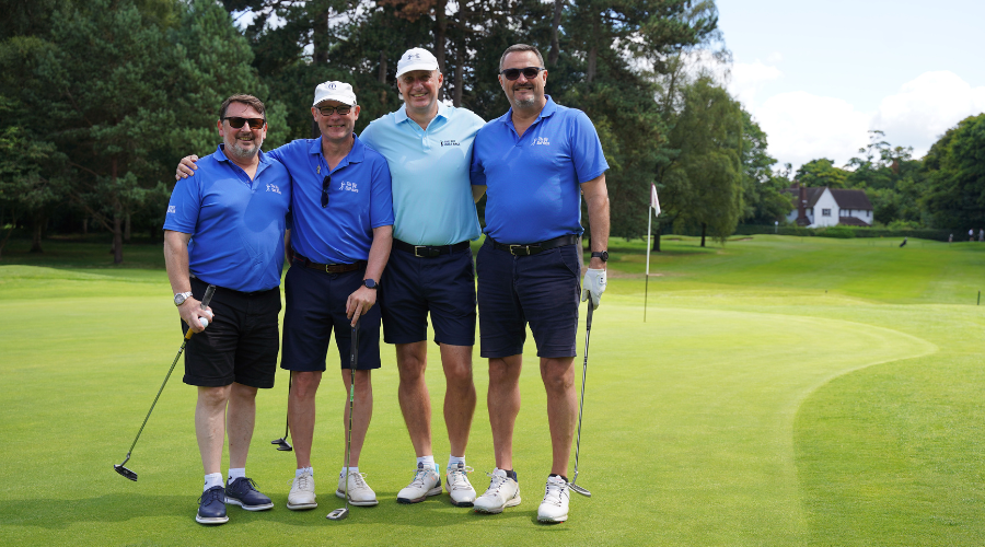 Four men standing on a golf course in front of the green, wearing blue shirts and caps