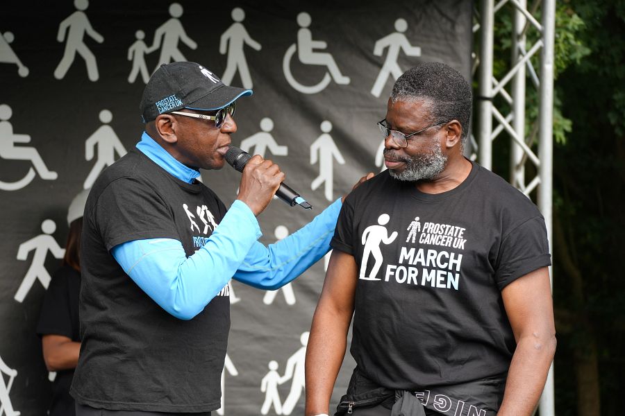 A man holding a microphone places his hand on a friend's shoulder and speaks to him on a stage at Prostate Cancer UK's March for Men event.
