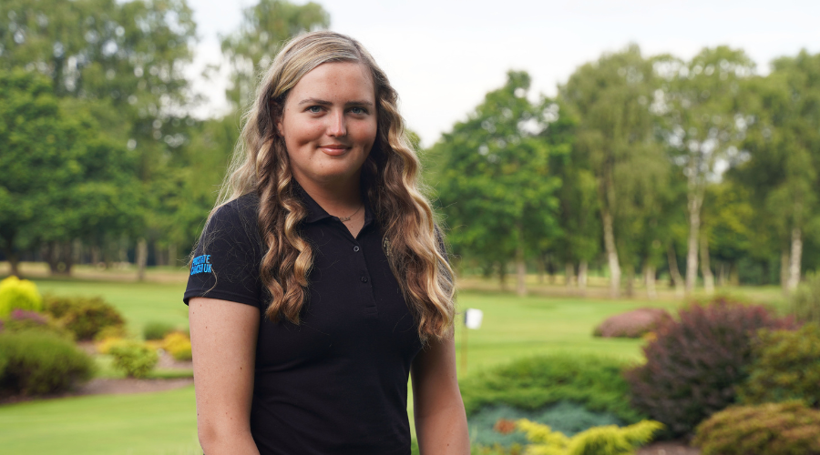 A young woman with blonde hair standing on a golf course, wearing black golfing kit and a Prostate Cancer UK Man of Men badge.