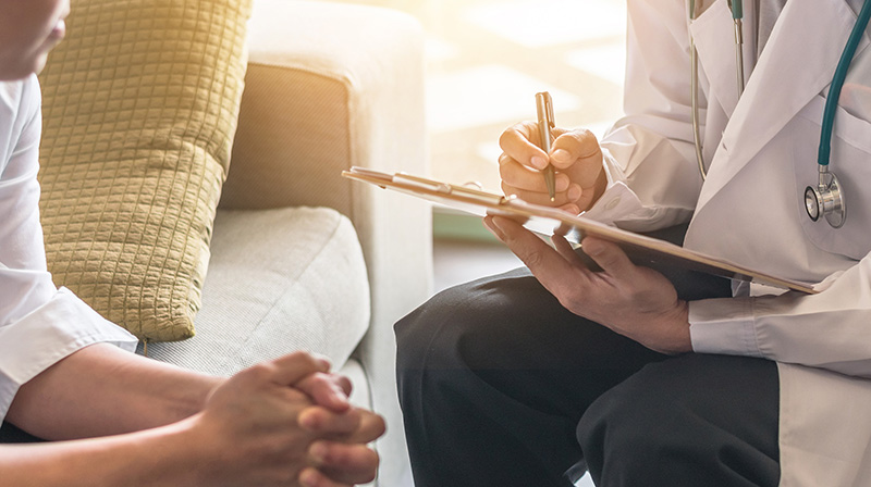 A man sitting on a sofa talking to a doctor who is writing on a clipboard