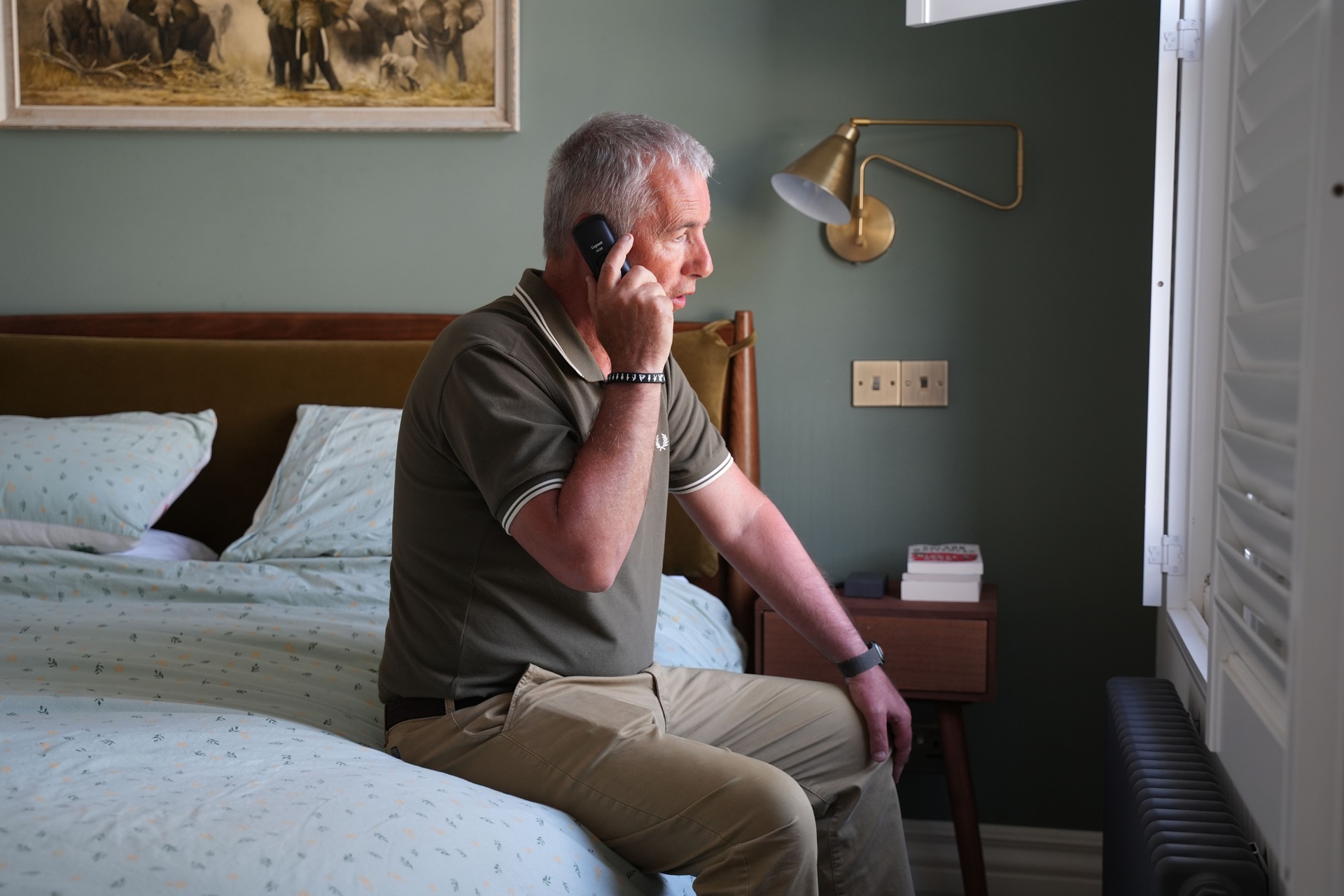 A man wearing a green shirt sitting on a bed and talking on the phone. 