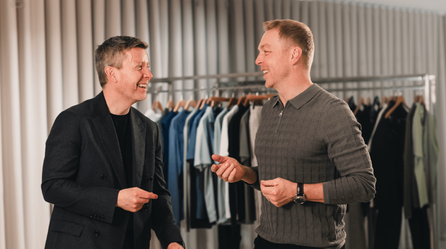 A man in a black jacket and a man in a grey sweater laughing and smiling in a men's clothing store